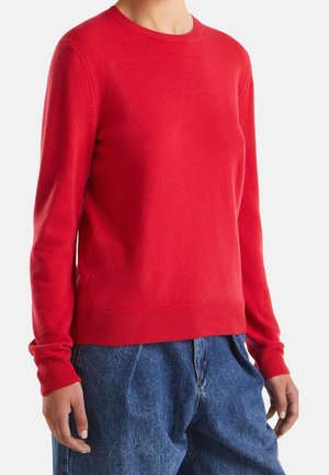 Nainen pukeutunut istuvaan punaiseen pitkähihnaiseen neulepaitaan ja väljääriin sinisiin farkkuihin, seisoo yksiväristä valkoista taustaa vasten.