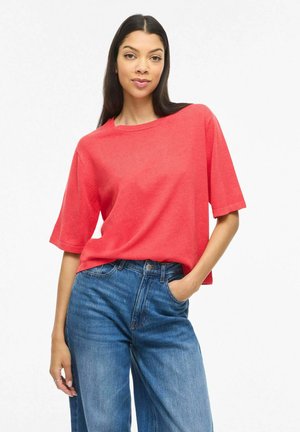 Femme debout, la main dans la poche, portant une chemise rouge à manches courtes et un jean taille haute bleu, sur fond blanc.