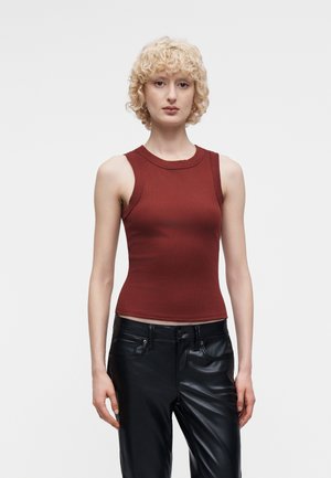 Femme aux cheveux blonds bouclés et courts portant un haut sans manches couleur rouille et un pantalon en cuir noir, debout devant un fond blanc.