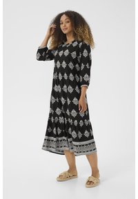 Schwarzes Kleid mit weißen Blumenmustern, Dreiviertelärmel und gestuftem Rock. Kombiniert mit beigen geflochtenen Slide-Sandalen.