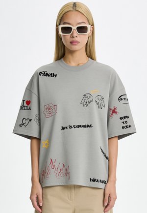 Person wearing gray oversized T-shirt with black and red doodles, text, angel wings, flames, and beige pants, paired with white rectangular sunglasses.