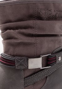 Bota de ante gris con una correa texturizada de color gris oscuro, hebilla de metal plateado y acentos en rojo y negro, que presenta costuras visibles y pliegues.