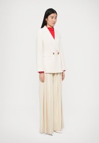 Witte dubbelgeknoopte blazer met gouden knopen, gedragen over een rode coltrui en crèmekleurige geplooide broek, gecombineerd met witte puntige schoenen.