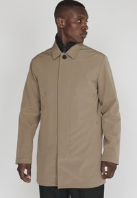 Abrigo beige, impermeable, con un alto cuello, cierre frontal con botones y bolsillos laterales. Textura suave, corte entallado y diseño minimalista.