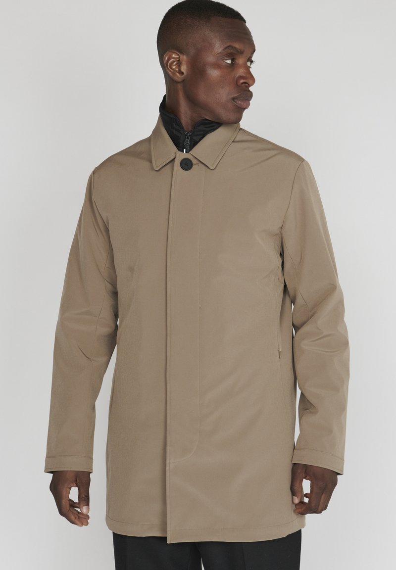 Abrigo beige, impermeable, con un alto cuello, cierre frontal con botones y bolsillos laterales. Textura suave, corte entallado y diseño minimalista.