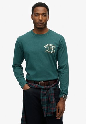 Teal langærmet t-shirt med "SUPERDRY JAPAN ATHLETIC" print i creme på brystet. Mørke jeans, brun bælte, ternet skjorte bundet om livet, ur på håndleddet.