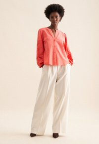 Koronkowa koszula z guzikami w kolorze koralowym, z haftowanymi rękawami w kształcie balonów, zestawiona z białymi spodniami o szerokich nogawkach i ciemnymi obcasami na neutralnym tle.