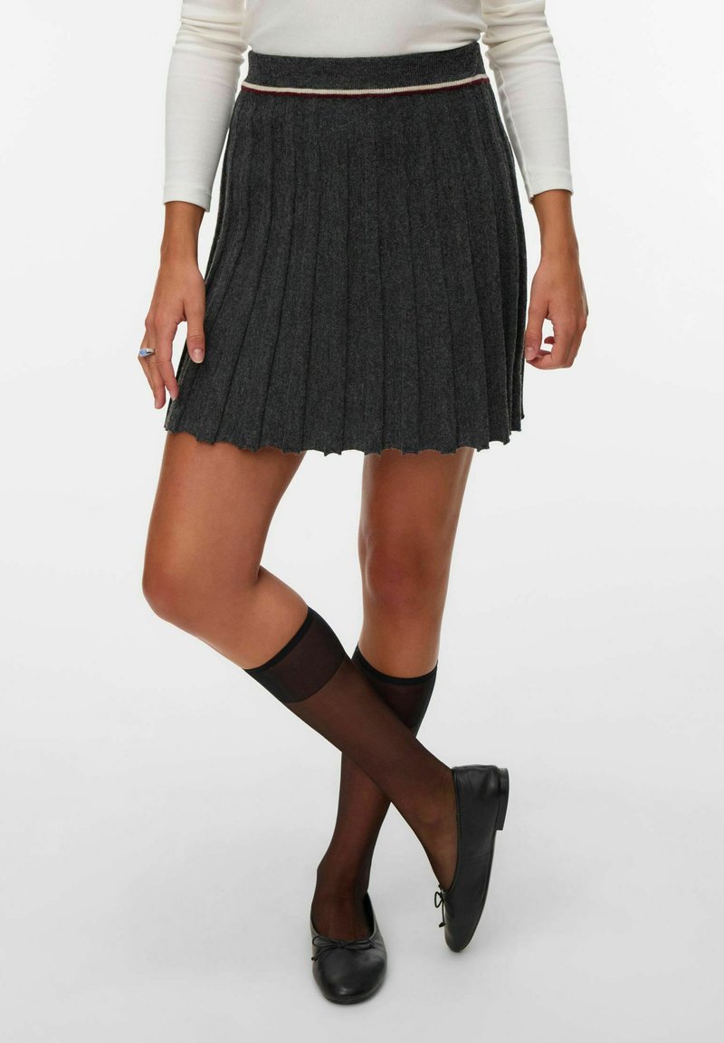 Femme portant une jupe plissée gris foncé, des chaussettes noires transparentes mi-mollet, des ballerines noires et un haut blanc à manches longues, debout les jambes croisées.