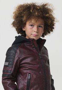 Bambino con capelli ricci che indossa una giacca in pelle bordeaux e nera con zip e cappuccio nero, che guarda leggermente di lato.