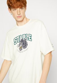 Bílý oversize tričko s grafikou zeleného nápisu "STAPLE" a černým designem vlka. Měkký bavlněný materiál, krátké rukávy, volnější střih.