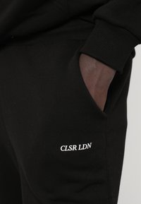 Czarne dresy z miękkiego materiału, z boczną kieszenią i białym napisem "CLSR LDN". Żebrowane mankiety przy kostkach. Subtelna tekstura widoczna.