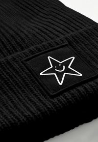 Black knitted beanie featuring a square patch with a white smiling star graphic. Textured fabric with a ribbed design.