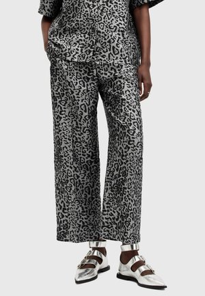 Personne portant un pantalon large imprimé léopard gris et noir et une chemise avec des chaussures plates argentées métalliques à boucle. Mains dans les poches.