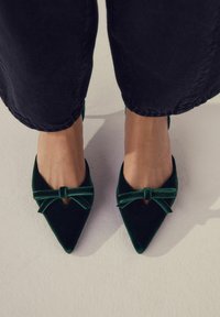 Picioare încălțate cu pantofi din catifea verde închis, cu detalii sub formă de fundă, asortați cu pantaloni negri scurți, pe un fundal deschis.