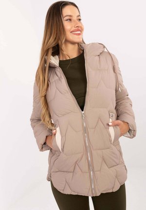 Smiling woman wearing beige quilted zip-up jacket with pockets, standing with hands in pockets, dressed in dark green shirt and pants.