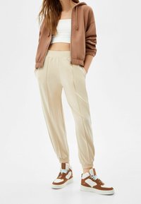 Kvinna iförd beige jogger-byxor, brun zip-up hoodie, vit crop top och bruna-och-vita högtoppsskor som står med händerna i fickorna.