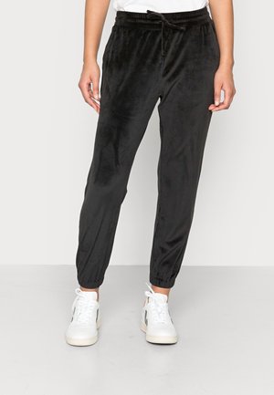 Person wearing black velour jogger pants with elastic cuffs and white sneakers, standing on a gray floor against a plain background.