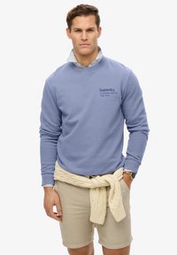 Sudadera azul claro de algodón, con puños y escote de canalé, y texto del logo en oscuro. Suéter de punto beige atado alrededor de la cintura.