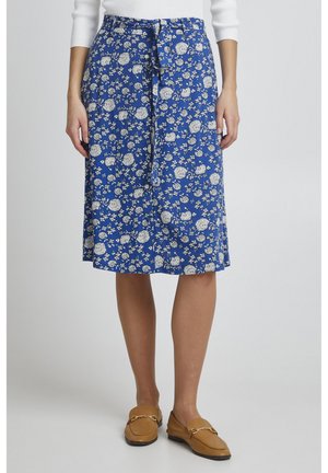 Jupe midi florale bleue avec des motifs de fleurs blanches, taille nouée, tissu fluide et ourlet légèrement évasé. Assortie avec des mocassins marron.