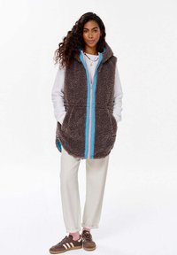 Chundelatá hnědá bezrukávová vesta s předním zipem, modrými a růžovými akcenty, bočními kapsami, nošená přes bílý top a krémové kalhoty.