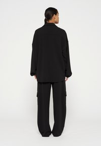 Schwarzes, übergroßes Hemd mit Kragen und überschnittenen Schultern, kombiniert mit schwarzen, weiten Cargo-Hosen mit zwei seitlichen Taschen.