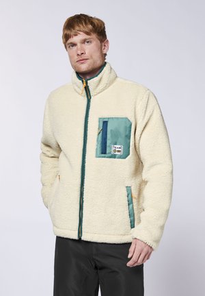 Man wearing cream fleece jacket with teal patch pocket and black pants standing against plain background.