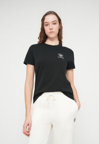 Μαύρο βαμβακερό μπλουζάκι με στρογγυλή λαιμόκοψη και κοντά μανίκια, με ένα μικρό λευκό γραφικό λογότυπο στην αριστερή πλευρά του στήθους.