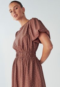 Smešasta haljina smeđe boje s točkicama, s kratkim nabranim rukavima, skupljenim po struku i mekanom teksturom, s malim bijelim točkicama raspoređenim po tkanini.