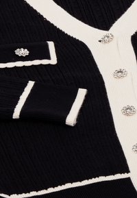 Schwarze und cremefarbene Strickjacke mit gerippter Struktur, V-Ausschnitt, dekorativen Kristallknöpfen und Kontraststreifen an den Bündchen und Taschen.