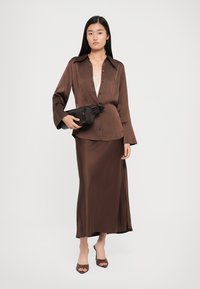Brown satin blouse with a pointed collar, paired with a matching long skirt. The model holds a black textured clutch and wears brown open-toe heels.