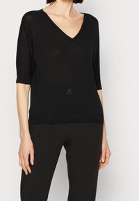 Zwarte gebreide trui met een V-hals, driekwartmouwen en een aangesloten zoom. Heeft een centrale accentknop en een lichte glans in de stof.
