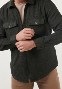 Camisa de cremallera de color verde oscuro y texturizada, con dos bolsillos frontales, detalles de botones y un dobladillo redondeado, llevada sobre una camiseta interior de color claro.