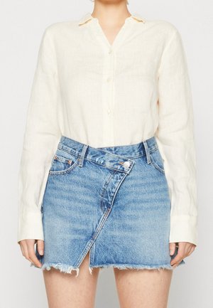 Femme portant une chemise blanche à manches longues boutonnée rentrée dans une mini-jupe en jean asymétrique bleue taille haute avec un ourlet effiloché.