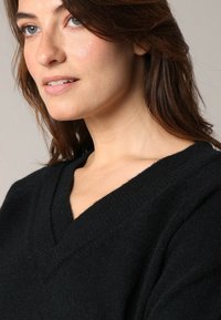 Femme à la peau claire et aux cheveux bruns portant un pull noir à col en V, regardant légèrement sur le côté avec une expression neutre.