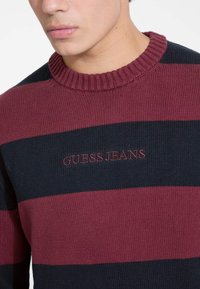 Brede gebreide trui met horizontale bordeauxrode en marineblauwe strepen, geribde ronde hals, en geborduurd "GUESS JEANS" logo op de borst.