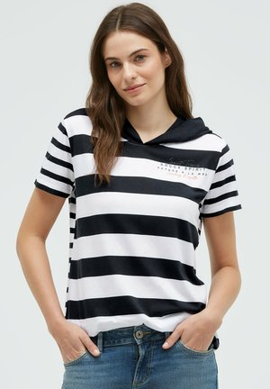 Young woman with long brown hair wearing a black-and-white striped short-sleeve hooded shirt and blue jeans, hands in pockets.