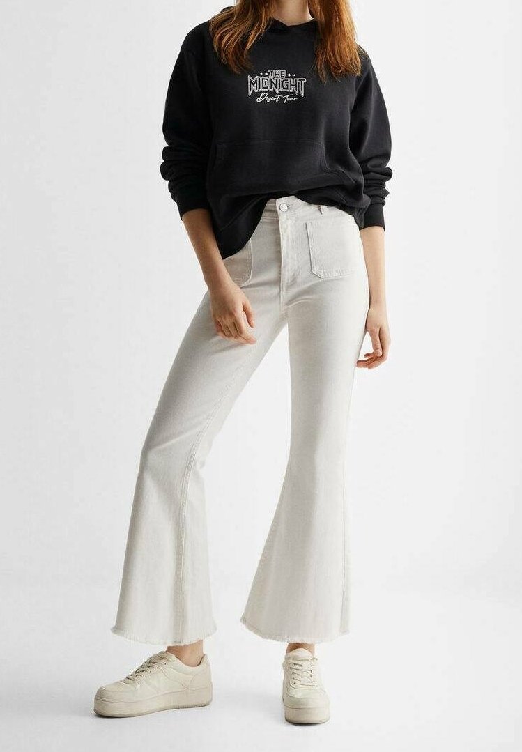 Femme portant un sweat-shirt noir avec le texte « The Midnight Desert Tour », un jean blanc évasé et des baskets à plateforme beige, debout devant un fond blanc.