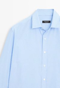 Chemise bleu clair à rayures verticales blanches, col à pointes et boutons blancs sur le devant, étalée à plat sur un fond blanc.