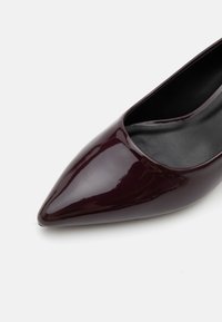 Chaussure à talon aiguilles brillante de couleur bordeaux foncé avec doublure intérieure noire sur fond blanc.