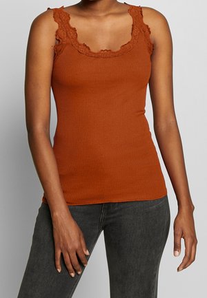 Vrouw draagt een mouwloze, geribbelde top in brandy oranje met kanten rand en donkere grijze jeans, staand tegen een effen grijze achtergrond.