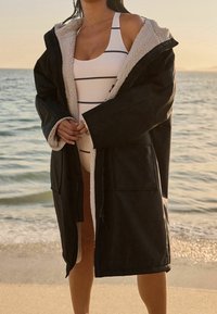 Zwarte buitenjas met een zachte roomkleurige voering, voorzien van een capuchon, oversized pasvorm en twee voorzakken, gedragen over een gestreept wit badpak.