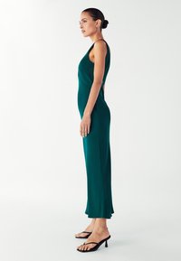 Dlouhé, těsně přiléhavé zelené šaty z hladkého materiálu s tenkými ramínky, které mají mírné rozšíření na spodním okraji, nošené s černými sandály na podpatku.