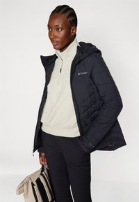 Svart dunjakke med hette, med kviltet design. Brukt over en lys beige fleece med half-zip, parret med svarte bukser og en stripete veske.