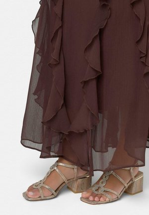 Pies con sandalias de tacón cuadrado beige con tiras bajo una falda marrón oscuro translúcida y fluida, con capas y bordes con volantes.