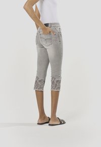 Szürke farmer capri nadrág, hímzett virágmintákkal a szegélyén, középmagas derékrésszel és hátsó zsebekkel, rojtos szegéllyel.