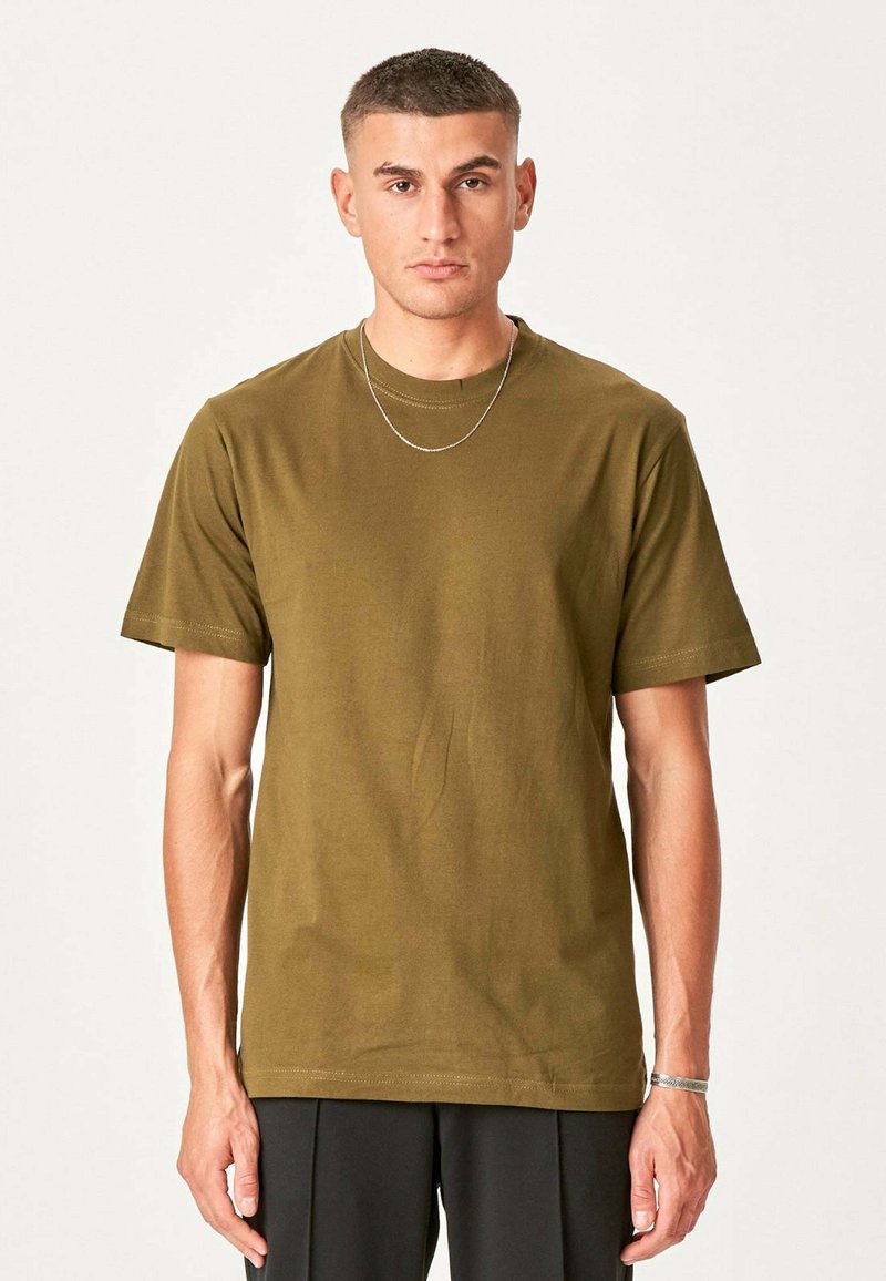 Mand med kort hår iført en olivengrøn t-shirt, sorte bukser, et tyndt halskæde og et armbånd, der står foran en ensfarvet baggrund.