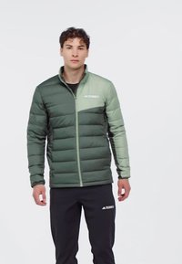 Giacca imbottita verde con colletto alto, dotata di un pannello verde più chiaro e logo sul petto, abbinata a pantaloni scuri.