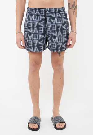 Uomo con pantaloncini da bagno neri e bianchi a motivo con loghi EA7, e sandali grigi abbinati EA7, in piedi contro uno sfondo bianco.