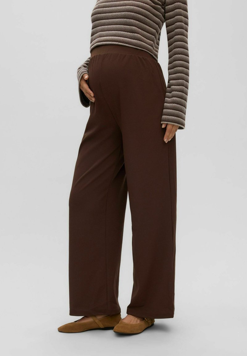 Personne enceinte portant un pantalon large marron, un haut à manches longues rayé et des chaussures plates marron, debout avec une main sur le ventre.