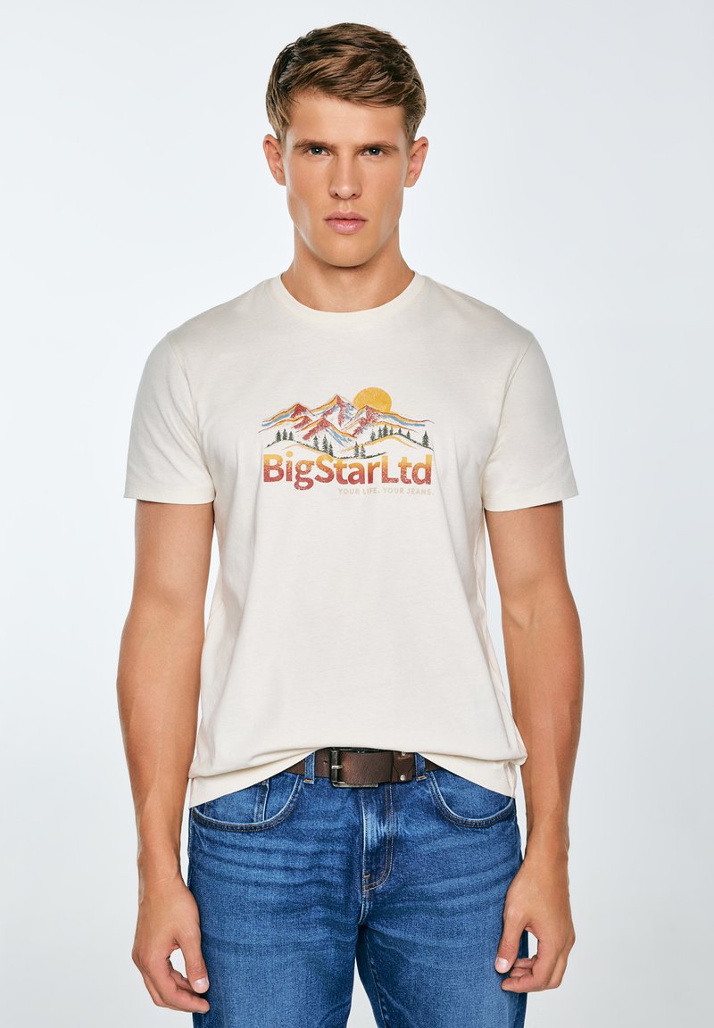 Krémové bavlněné tričko s barevným potiskem hory a slunce a textem "BigStarLtd"; doplněné modrými džíny a hnědým páskem.